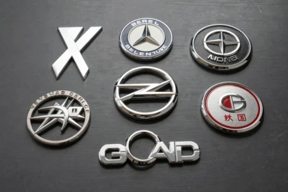 Gros plan de plusieurs logos de voitures en chrome et métal disposés sur une surface en acier brossé sombre, révélant les détails sculptés et la symbolique des emblèmes automobiles